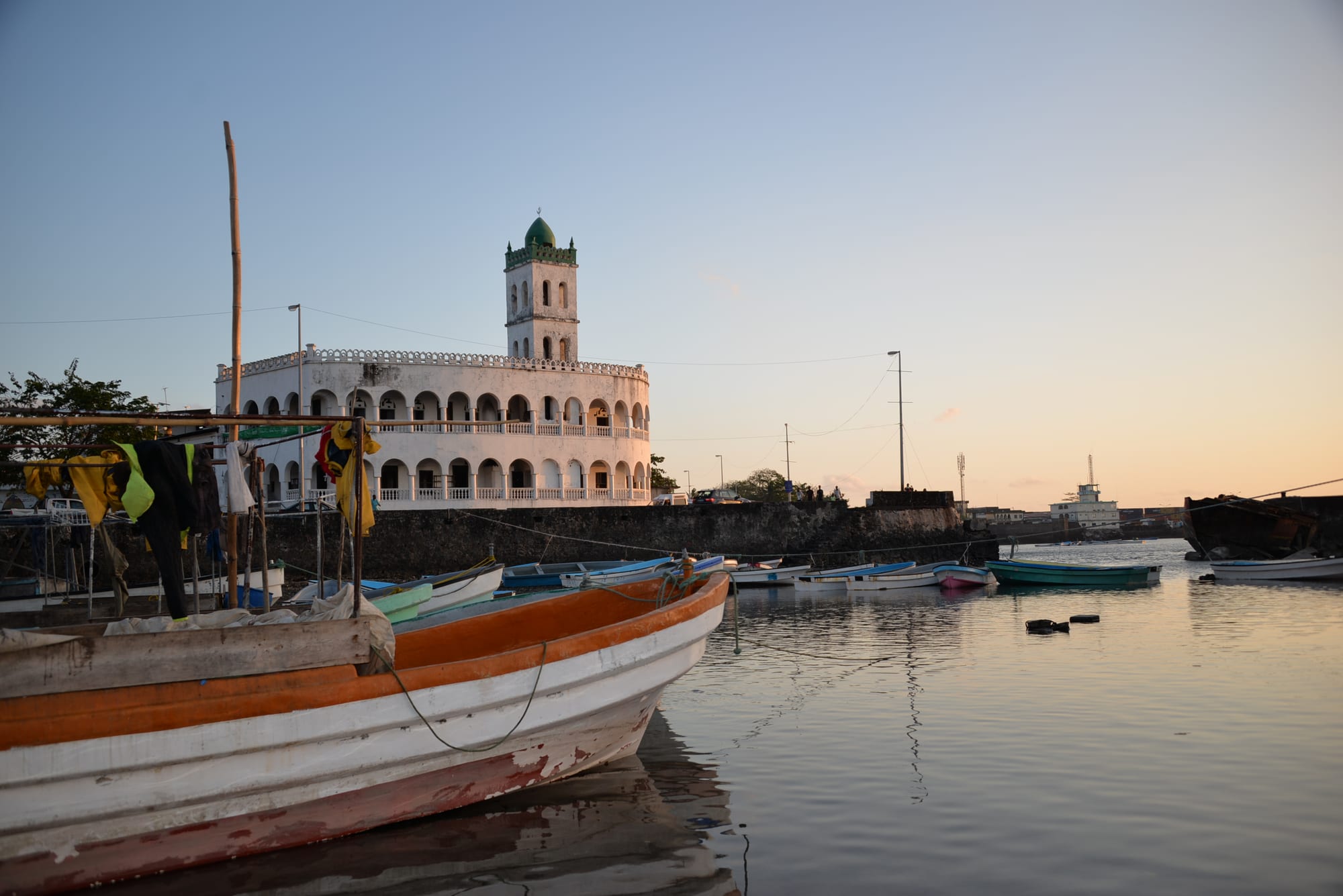A white two-storey mosque with arched arcades and a square minaret topped by a green dome, standing beside a harbour with small boats moored in calm water at dusk