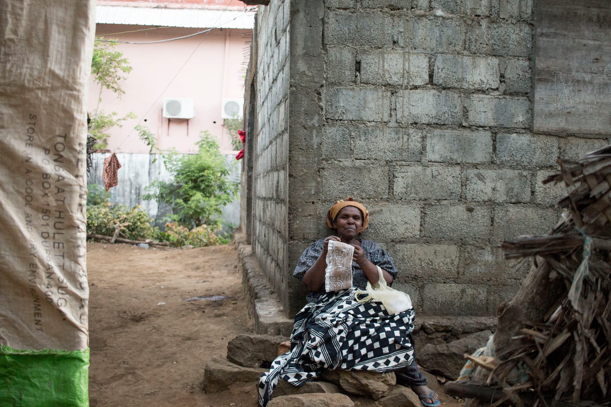 A woman in a headscarf and a black and white patterned wrap sits on a stone step against a breeze-block wall, holding up a white cloth cap she is embroidering by hand
