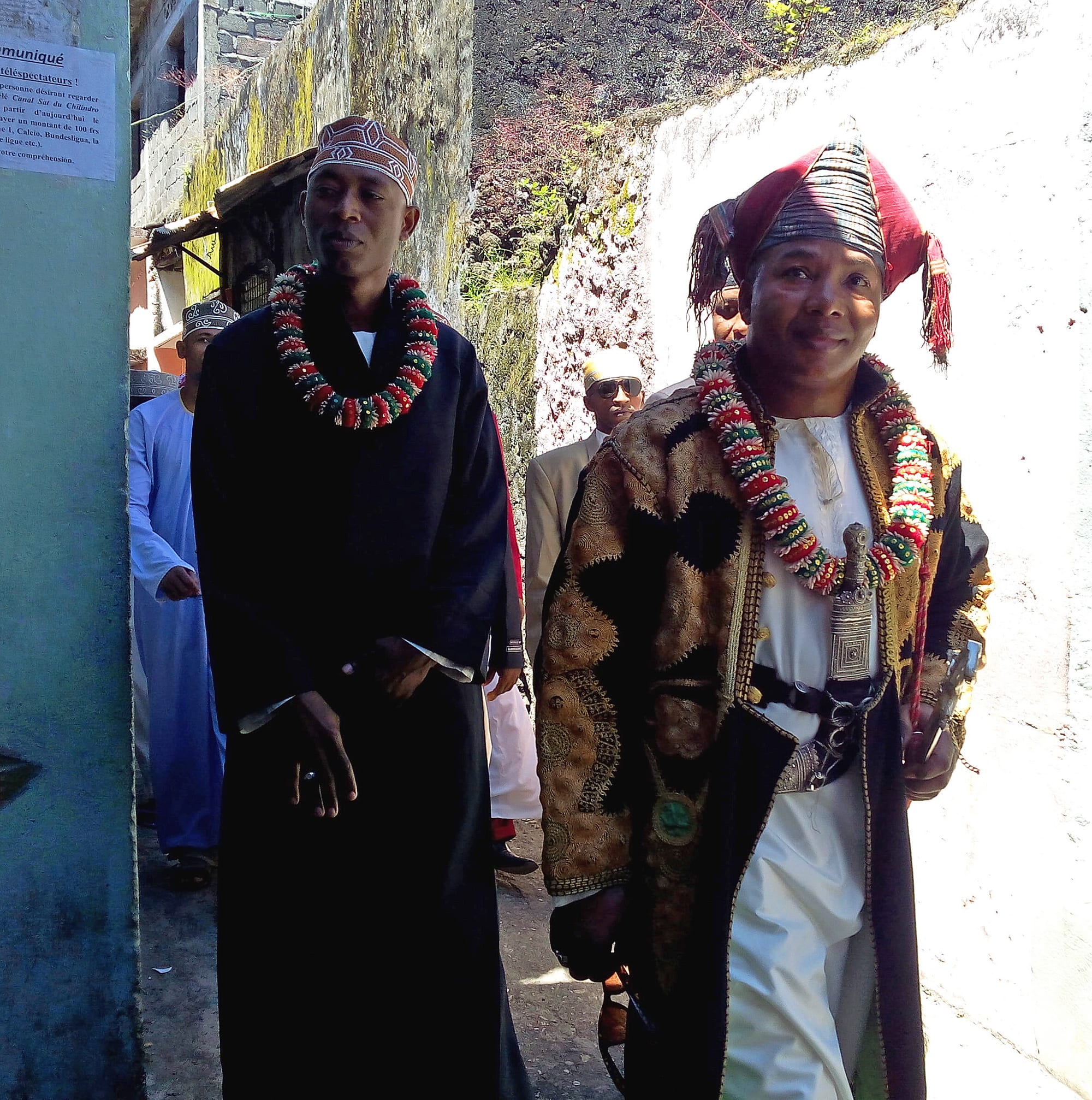 Two men in ceremonial dress walk along a narrow lane, one in a long dark robe and embroidered cap, the other in a gold-embroidered coat and red turban with a dagger at his belt, both wearing thick garlands