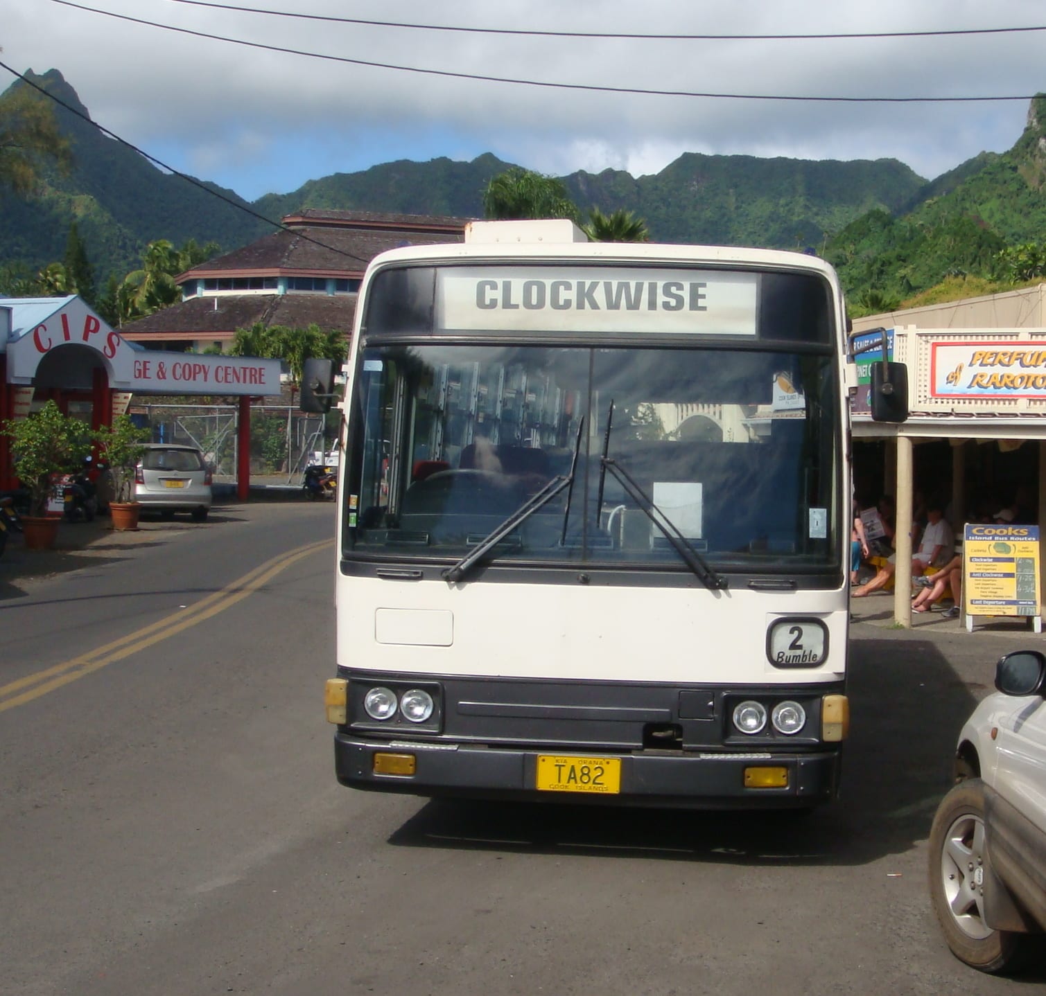 A white bus with CLOCKWISE displayed above the windscreen, parked on a street in Avarua with a bus-stop timetable sign visible