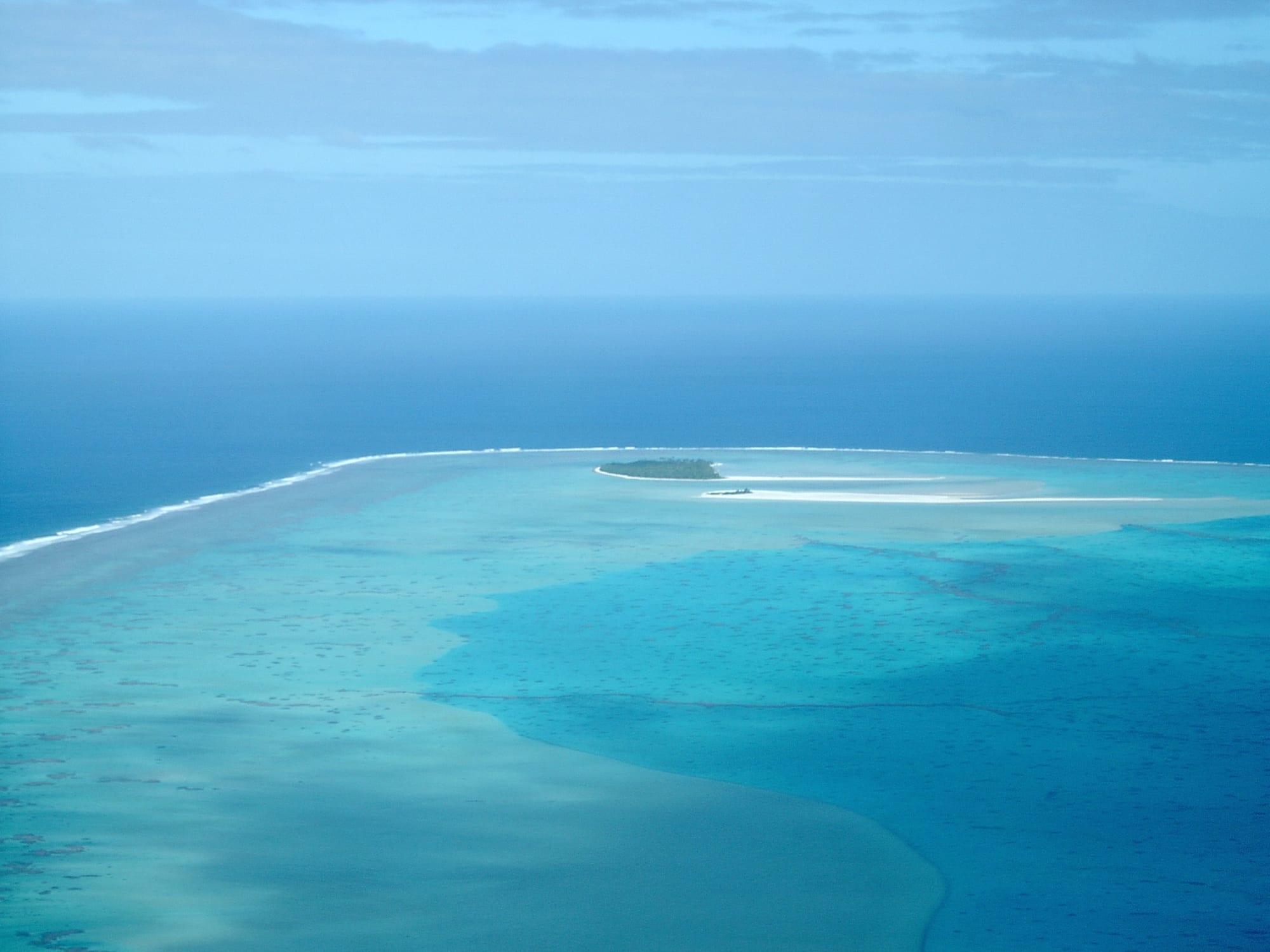 A small palm-covered islet with a broad white sandbank running out into shallow blue water