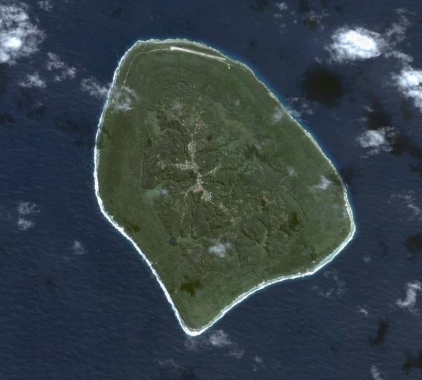 An aerial view of a roughly circular, densely forested island ringed by a pale reef edge