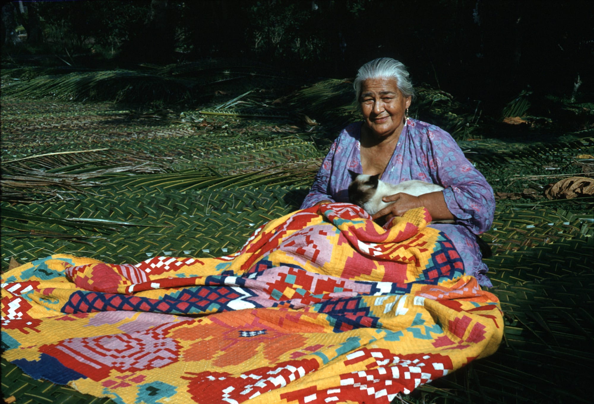 An older woman in a floral dress sits on a woven coconut-frond mat with a cat on her lap and a large, brightly coloured patchwork quilt spread across her knees