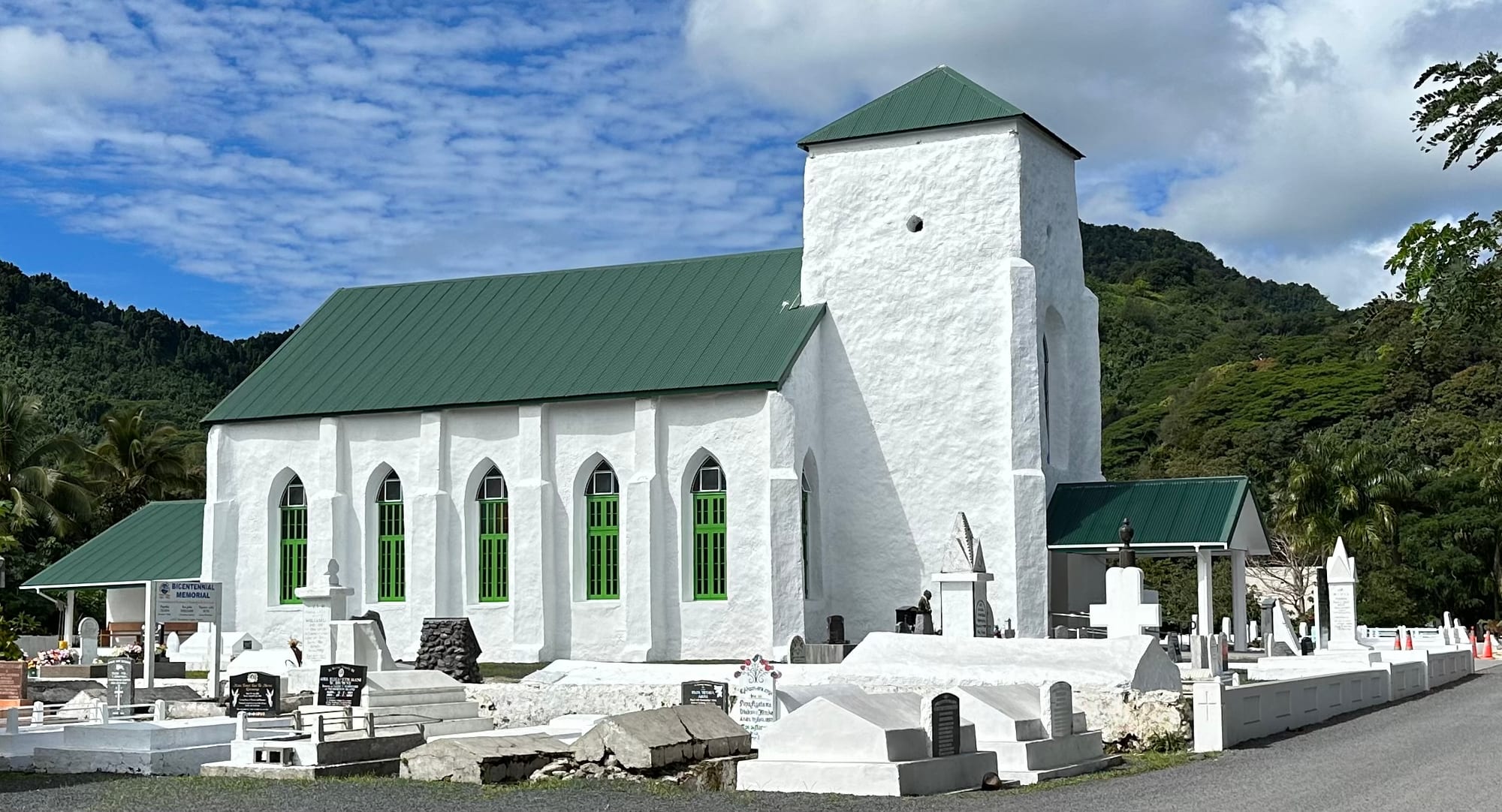 A whitewashed church with a square tower, green roof and green arched windows, surrounded by white-painted graves and memorials, with forested hills behind
