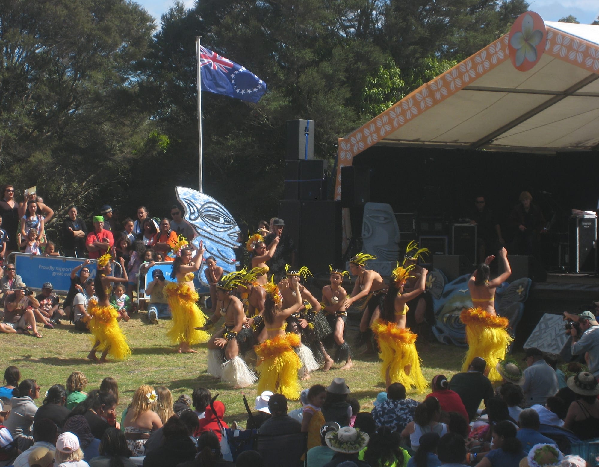 Women dancers in yellow grass skirts and feathered headdresses and men in white fringed skirts perform on a lawn in front of a seated crowd, with the Cook Islands flag flying above