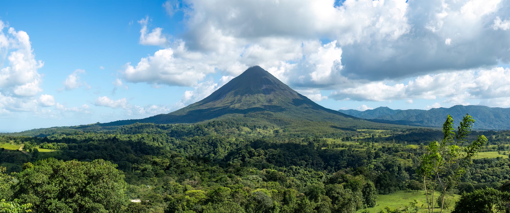 A symmetrical forested volcanic cone rising above green farmland under a partly clouded sky