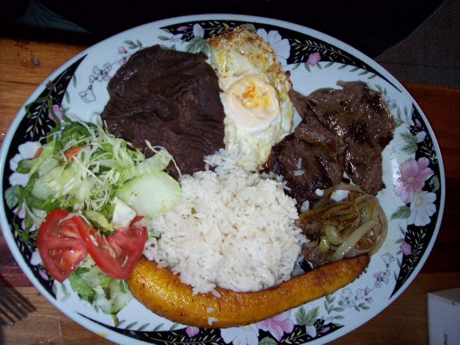 A plate divided into sections holding rice, plantain, meat, egg, beans and a cabbage salad