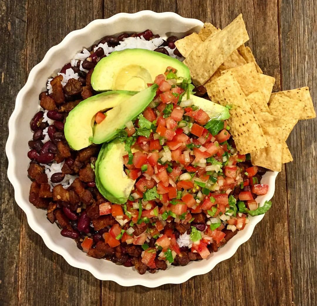 A bowl layered with rice, beans, fried pork pieces, avocado and tomato salsa