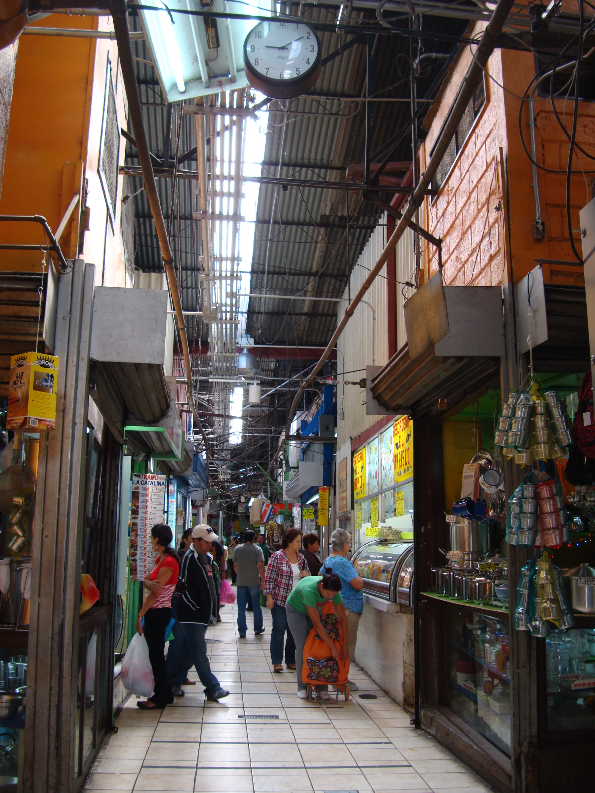 A narrow indoor market aisle in San José lined with small shop stalls, a large clock overhead, and shoppers walking through