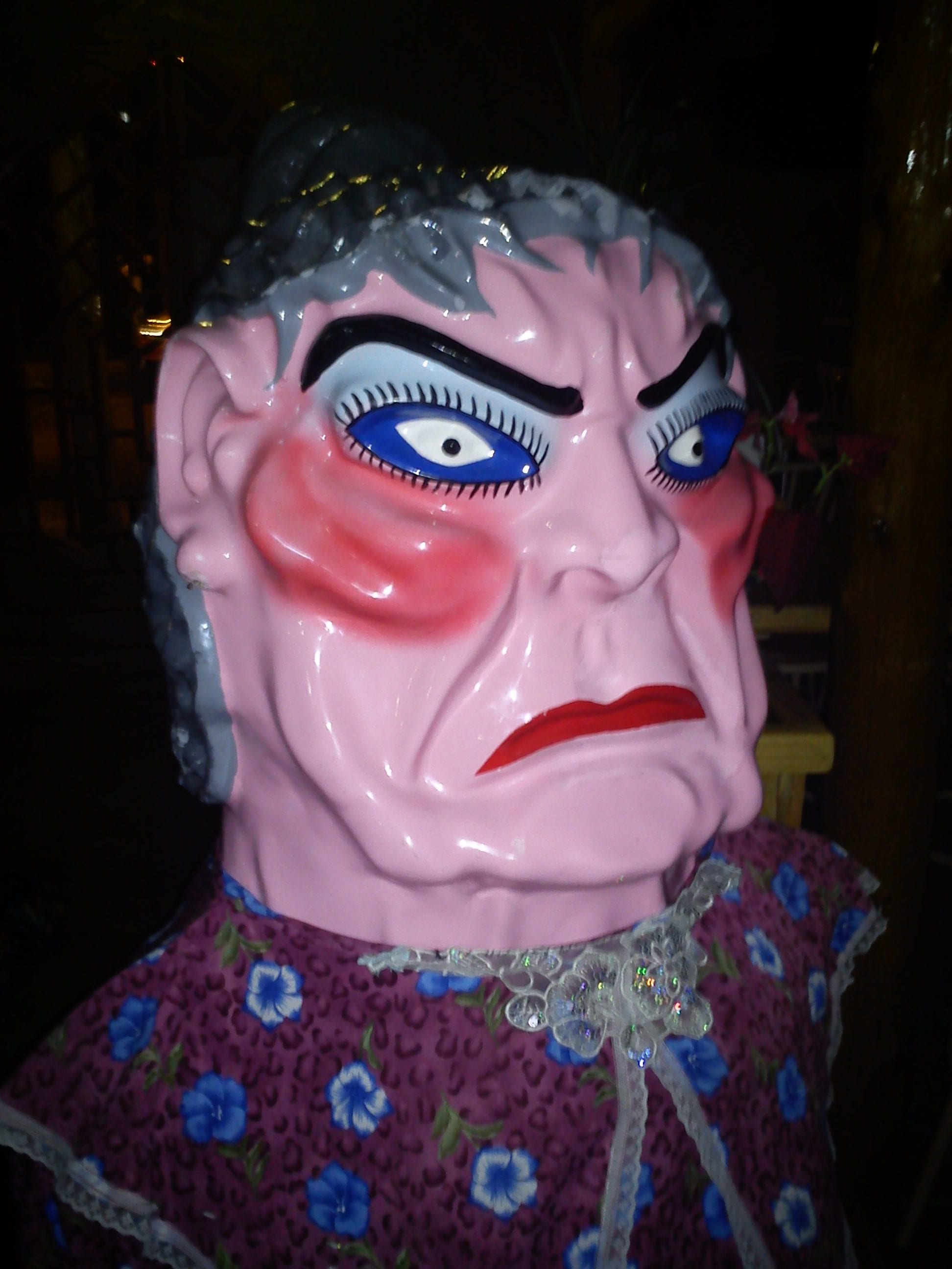 A close view of a large painted mascarada mask of a stern older woman with grey hair, heavy blue eye make-up and red lips, above a flowered dress