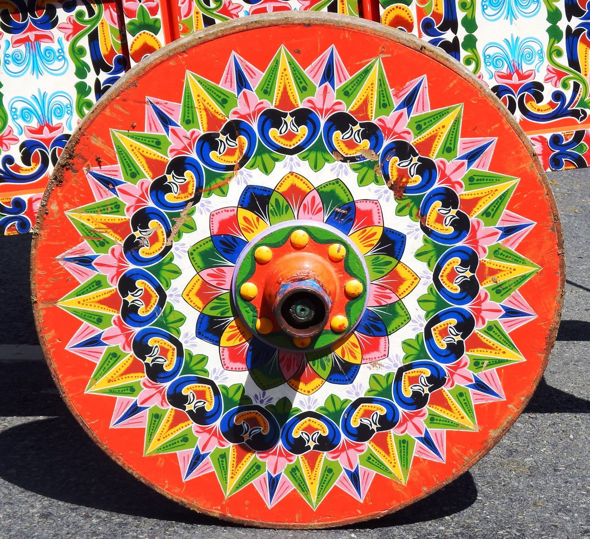 A solid wooden cartwheel painted bright orange with a radiating pattern of green, pink, yellow, blue and black star points and swirls around a central hub