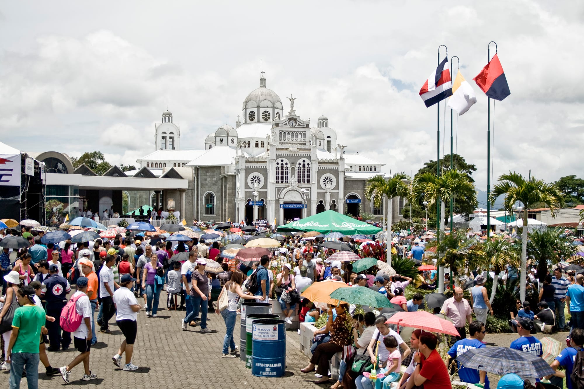 Large crowds with umbrellas fill a paved square in front of a grey-and-white domed basilica, with Costa Rican and Vatican flags flying on tall poles