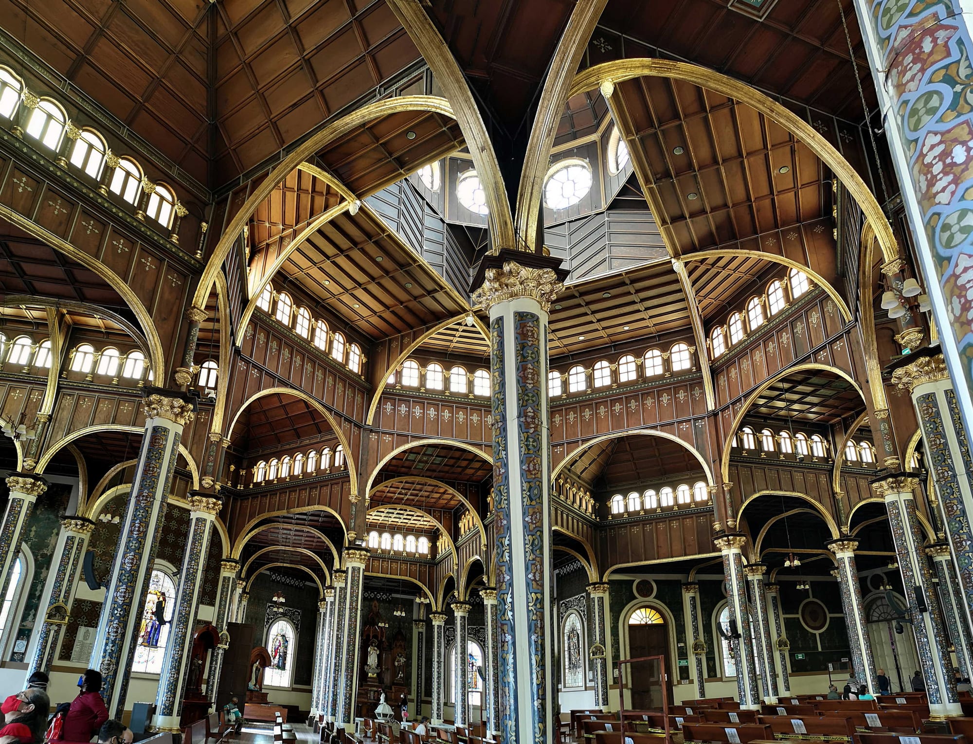 The interior of a large church with a timber ceiling, gilded arches and tall painted columns, with rows of wooden pews below