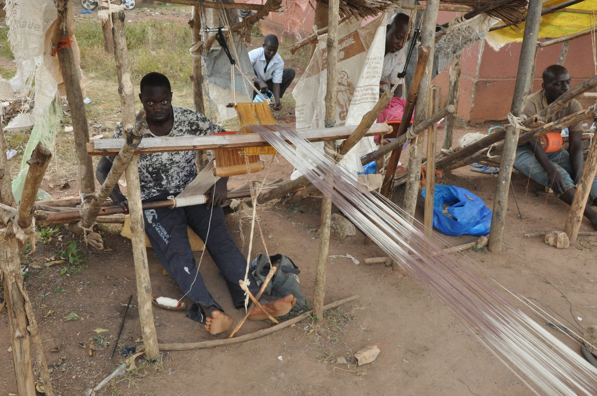 A man sits at a wooden hand loom set on posts outdoors, working a narrow strip of cloth, with long white warp threads stretched out in front of him and other weavers behind