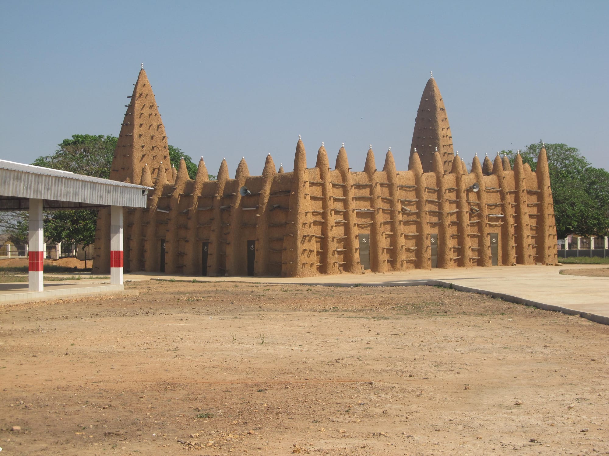 A long earthen mosque with rows of pointed buttresses studded with projecting wooden beams and two tall tapering towers, under a clear sky