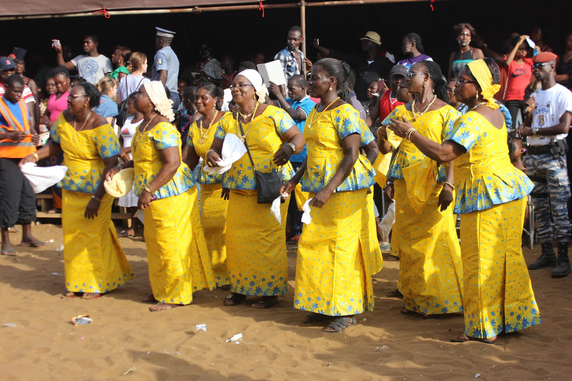 A line of women in matching yellow dresses with blue patterns and white headscarves dance on sand, holding white handkerchiefs, with a crowd watching behind
