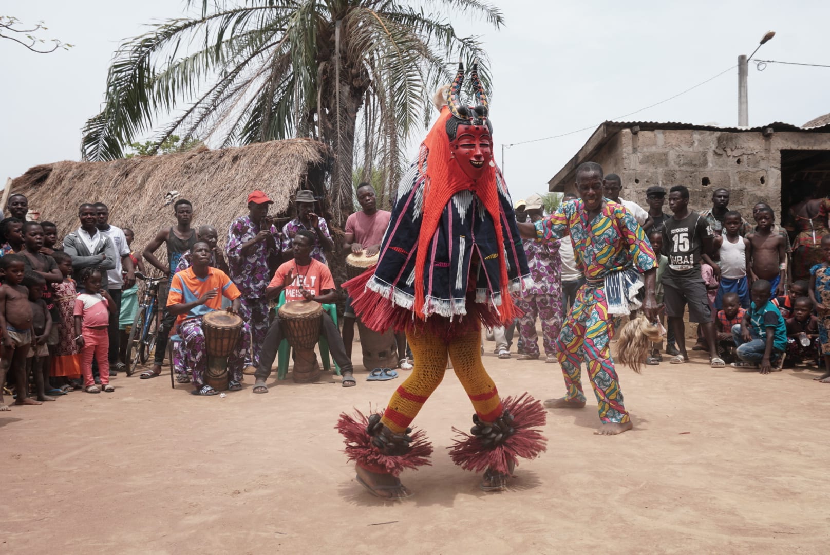 A masked dancer in a red and black costume with fringed ankle rattles dances on bare earth in a village, with drummers seated behind and a crowd watching