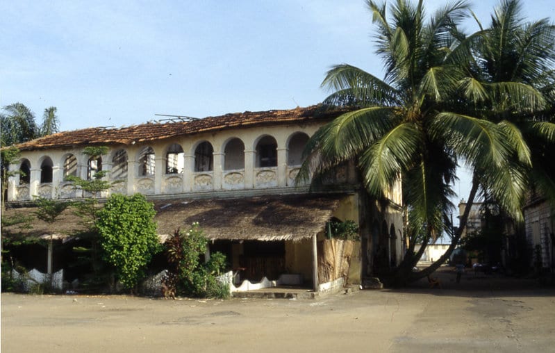 A grand but faded colonial building with an arched veranda, a palm tree beside it