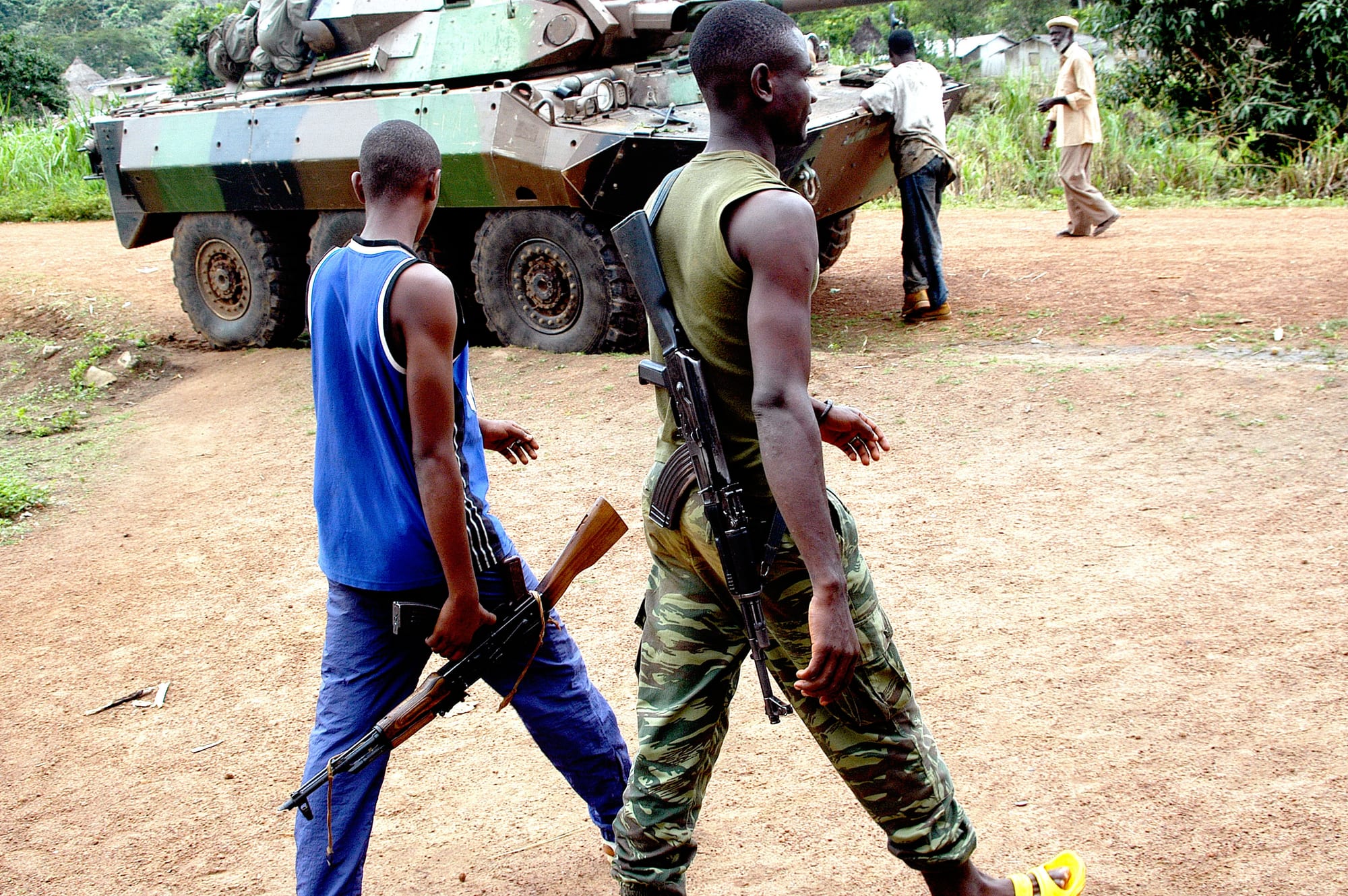 Two men carrying assault rifles, one in a blue vest and one in camouflage trousers, walk along a red-dirt village road past a camouflaged French armoured car