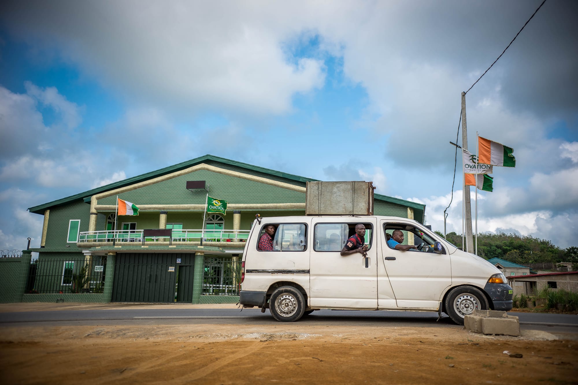 A white minibus with passengers leaning out of the open windows, driving past a green building with Ivorian flags
