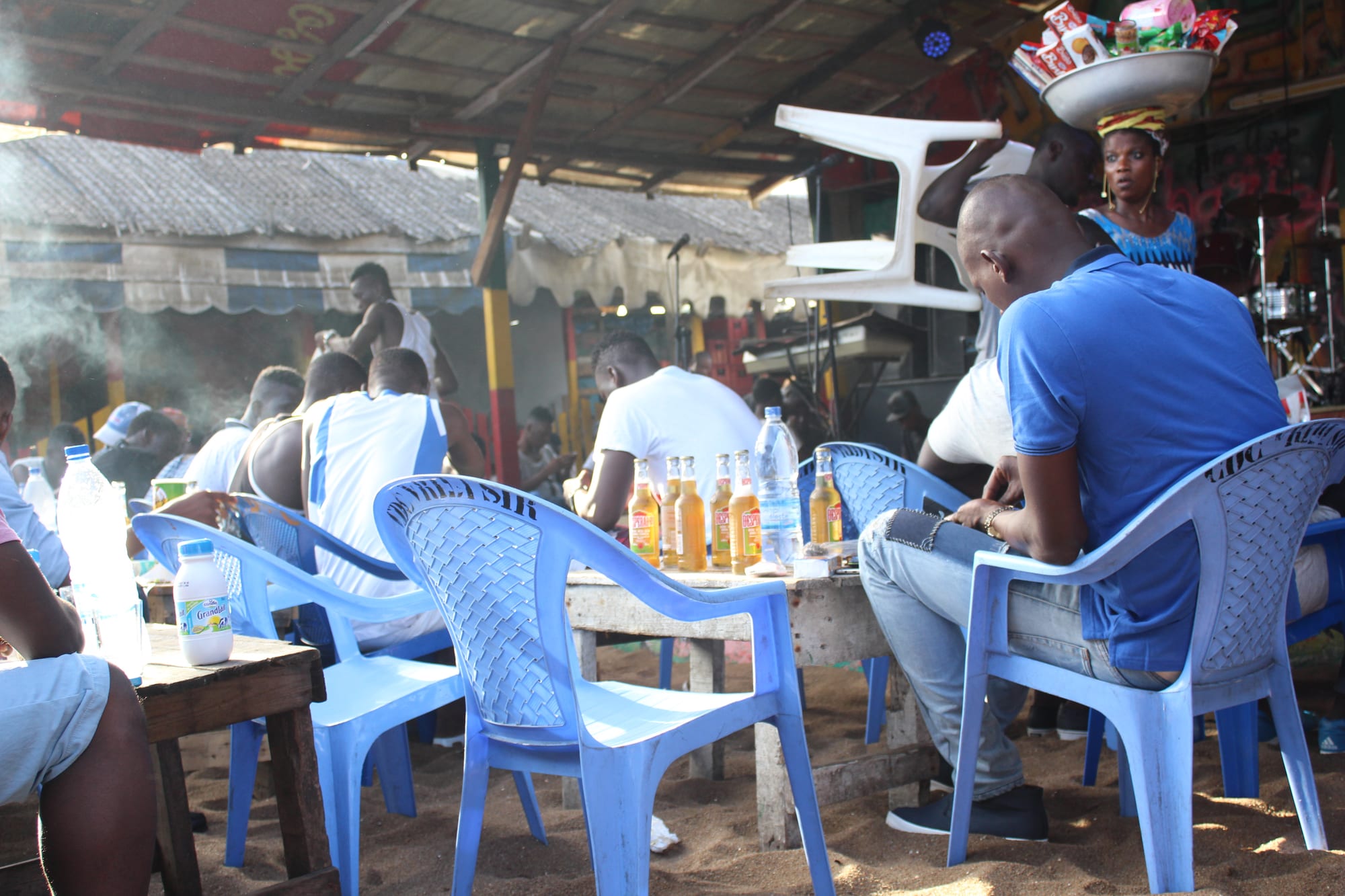 People sitting on plastic chairs at outdoor tables in a covered open-air eatery in Abidjan, with drinks on the tables and a live band playing behind