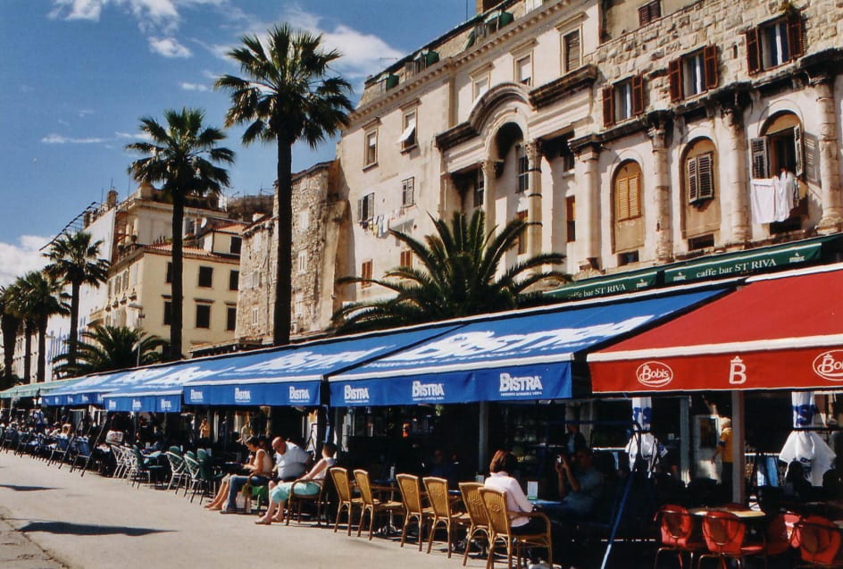 A row of waterfront cafes under blue and red awnings, with people seated at outdoor tables along a palm-lined promenade