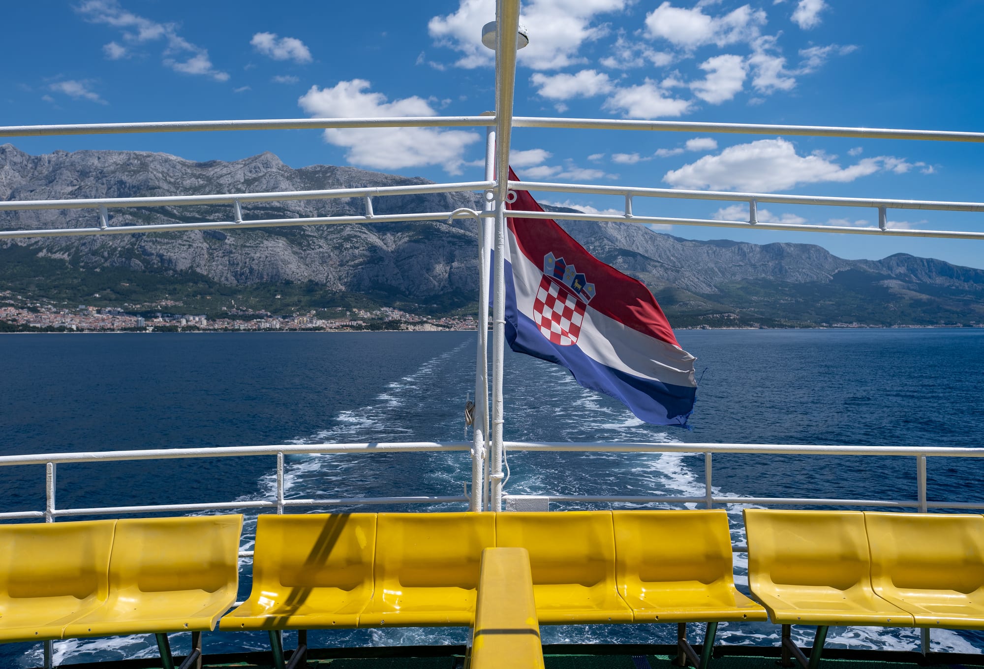 A Croatian flag flying from the stern of a ferry crossing open water, with mountains and a coastal town visible behind