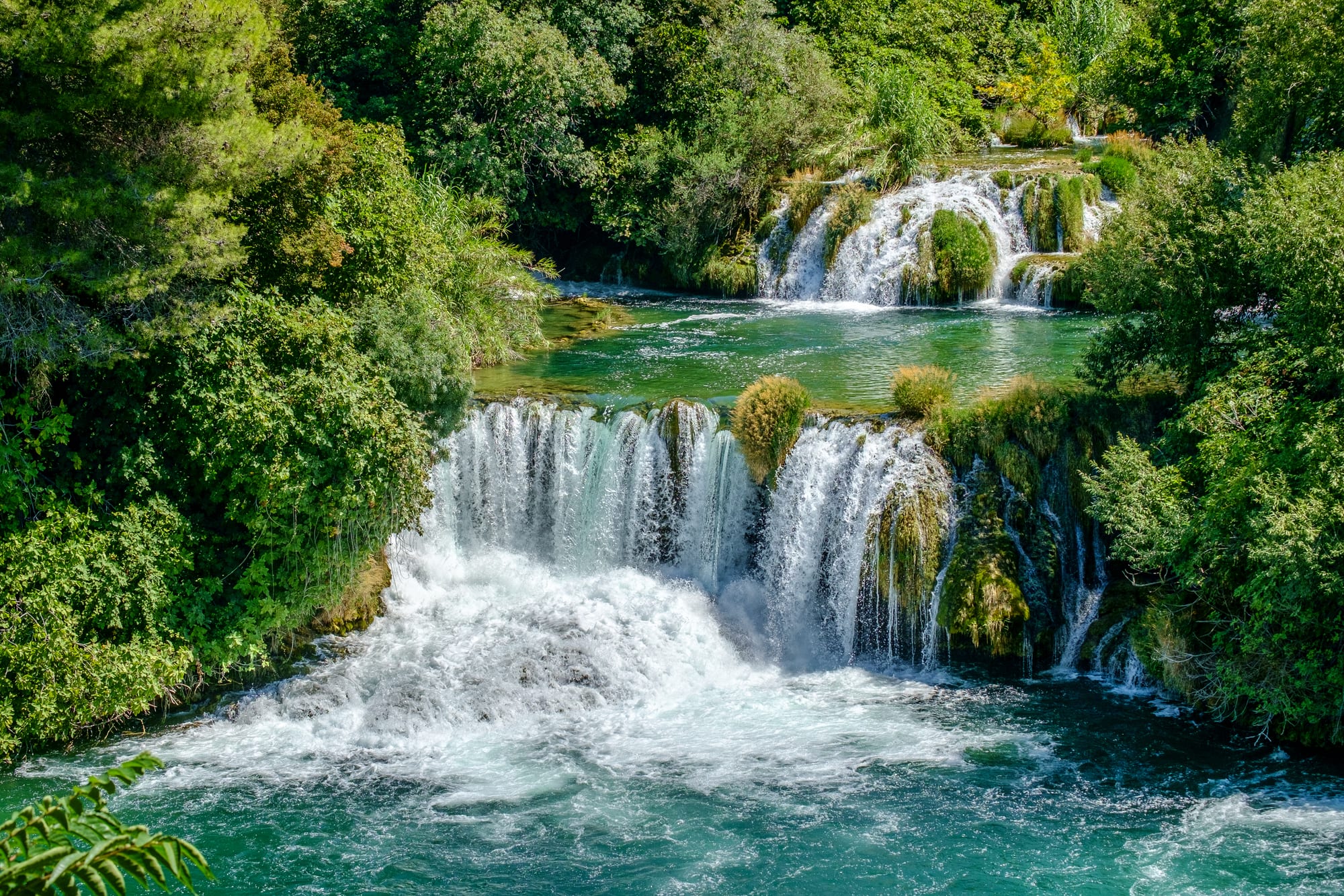 A wide set of stepped waterfalls tumbling over green travertine terraces into a river