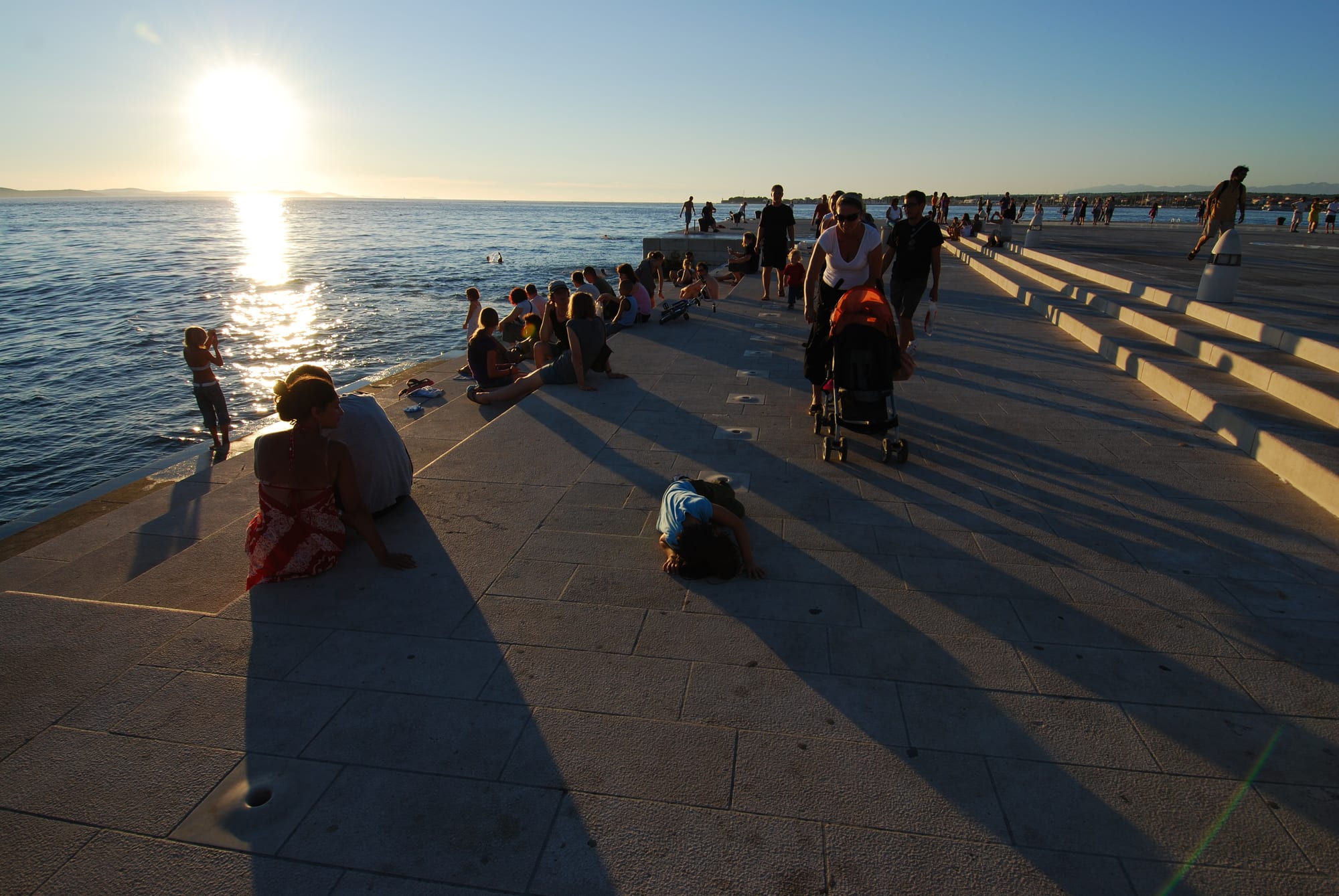 People sitting on wide stone steps by the sea at sunset, waves entering slots below