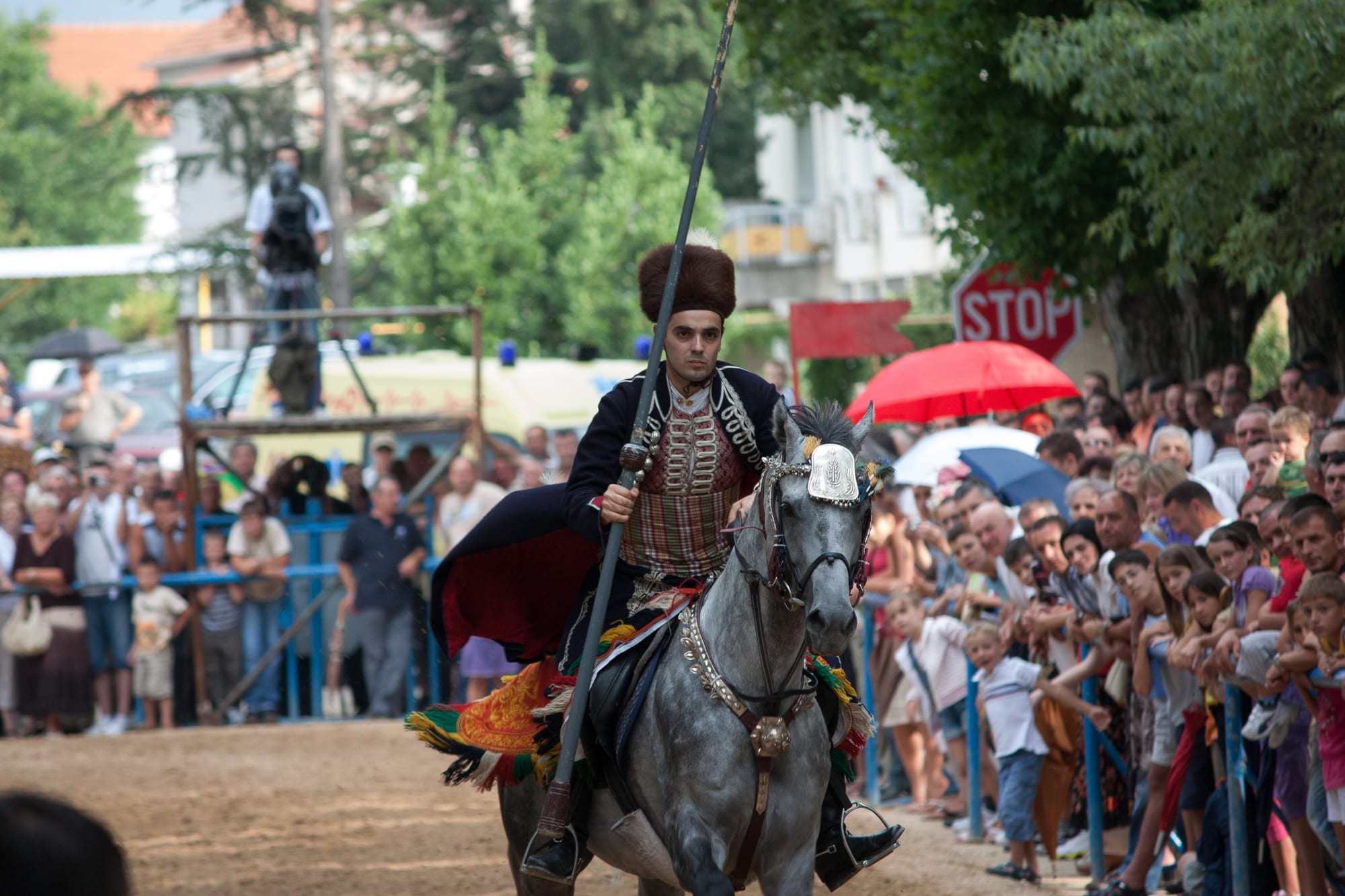 A rider in a fur hat, embroidered waistcoat and red-lined cloak gallops a grey horse along a dirt track holding a long lance, with crowds lining the barrier