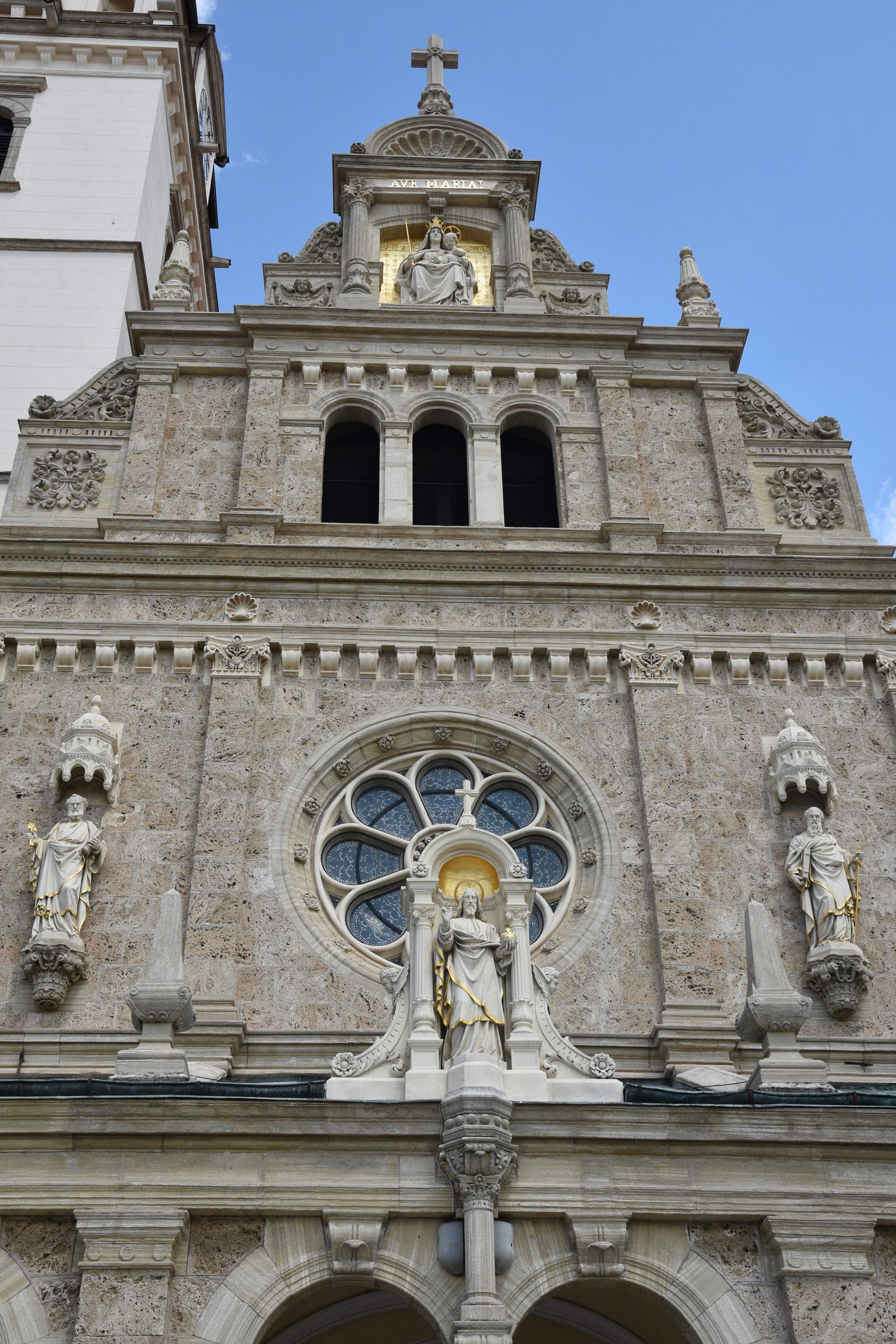 The pale stone facade of a basilica with a rose window, a statue of Christ above the entrance, saints in niches and a gilded statue of the Virgin and Child near the top