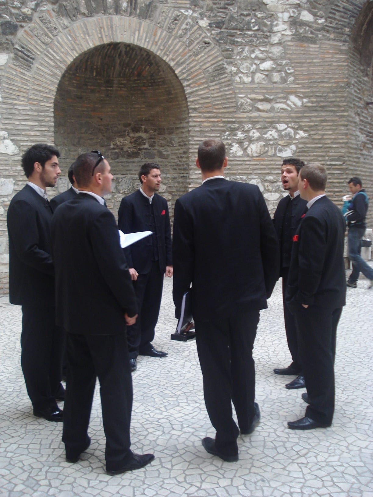 A group of men in dark suits with red pocket squares stand in a loose circle singing under a high stone and brick arch