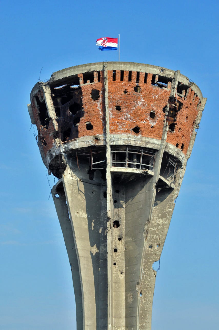 A tall concrete water tower riddled with shell holes, left standing above a rebuilt town