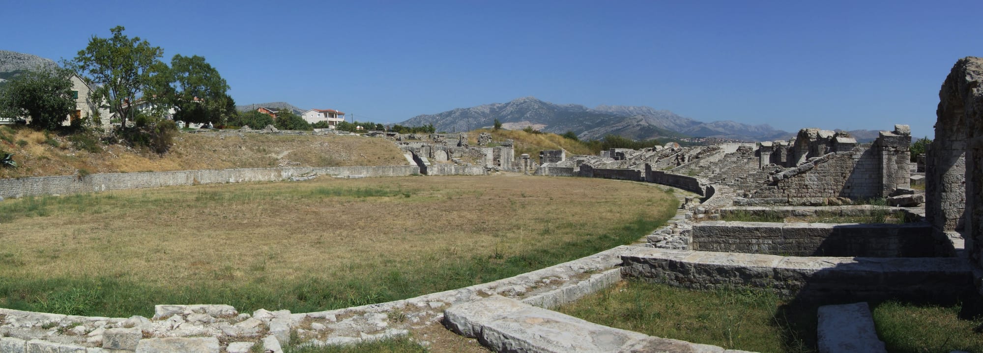 The low, curving stone foundations of a ruined Roman amphitheatre around a grassy arena, with mountains in the distance