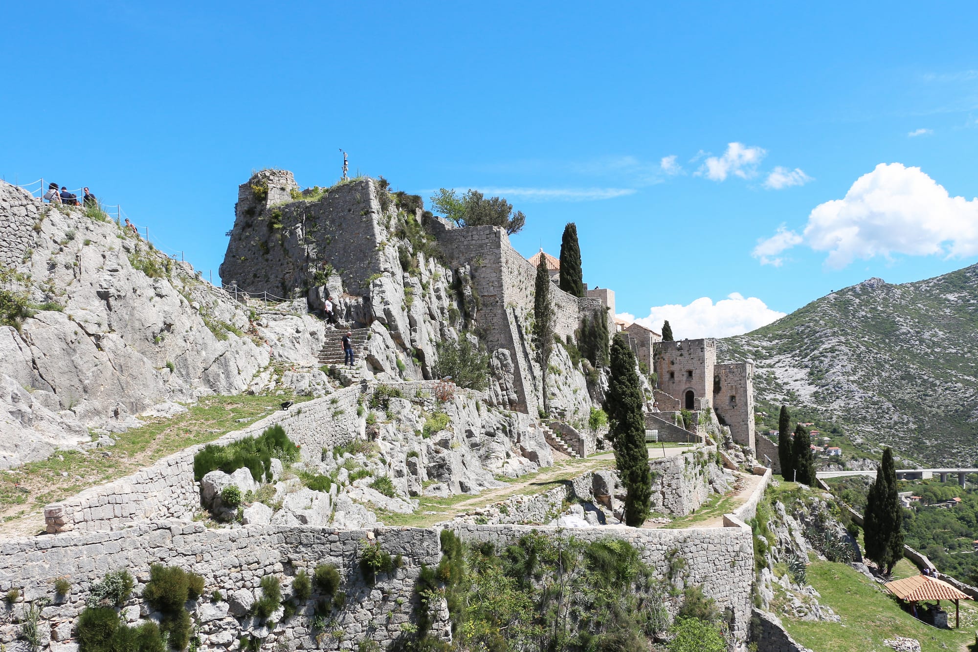 A stone fortress of walls, towers and stairways built along the crest of a bare limestone ridge, with rocky hills behind under a blue sky