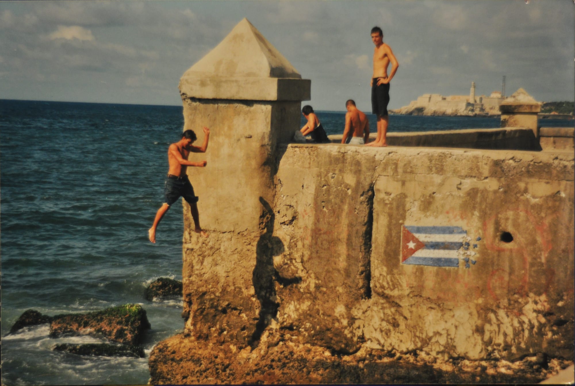 Young men jumping and standing on the stone Malecon seawall in Havana above the sea, with a fortress visible across the bay