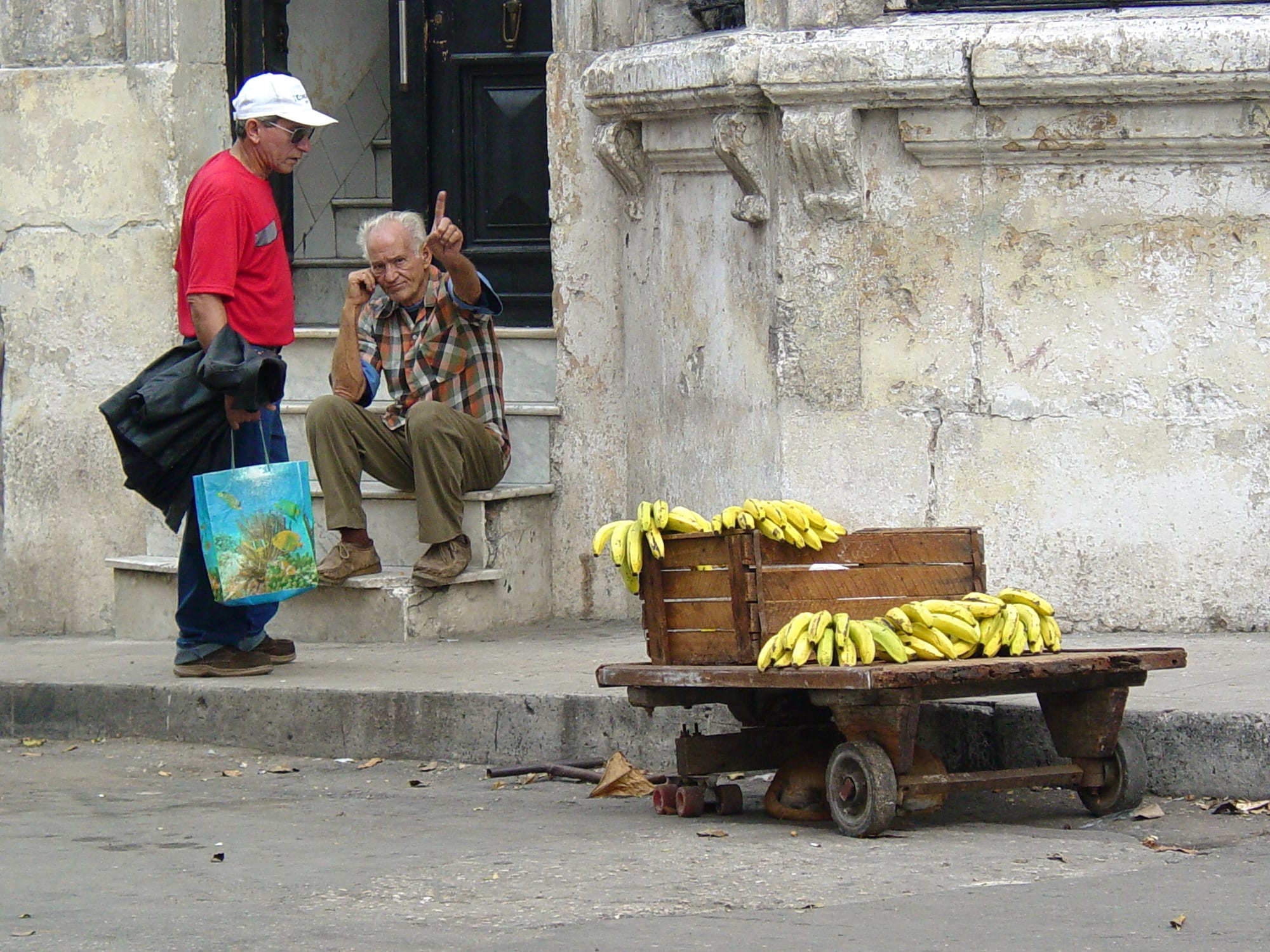 A street vendor's wooden cart piled with bunches of bananas on a Havana street, with two men talking on a nearby doorstep
