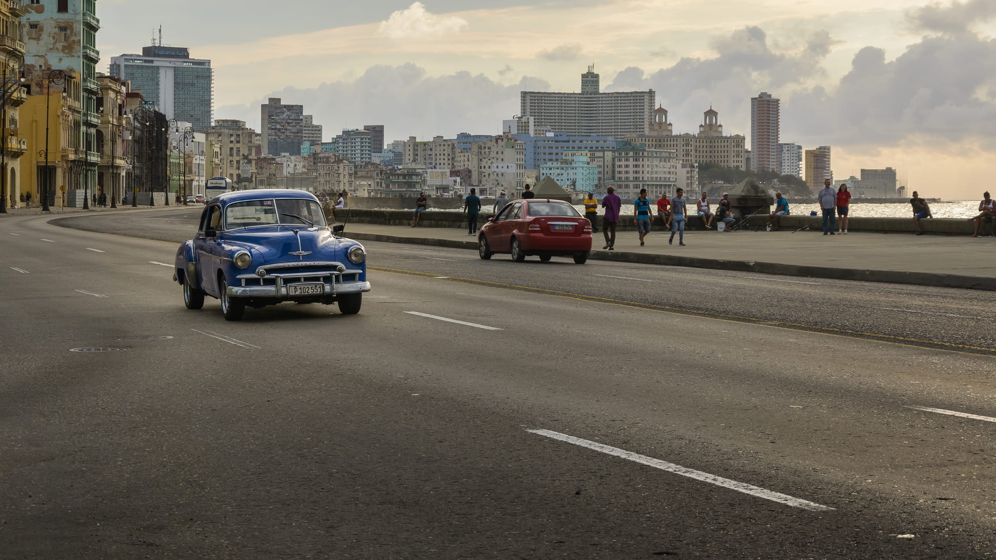A vintage blue American car driving along a seafront road with waves breaking over the wall