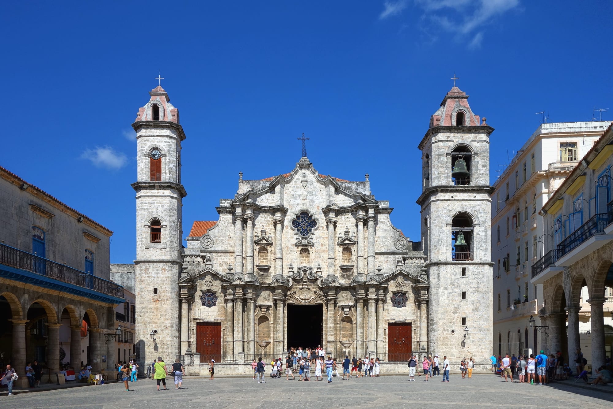 The pale stone baroque facade of Havana Cathedral with a curving central gable and two unequal bell towers, above a paved square with visitors under a deep blue sky