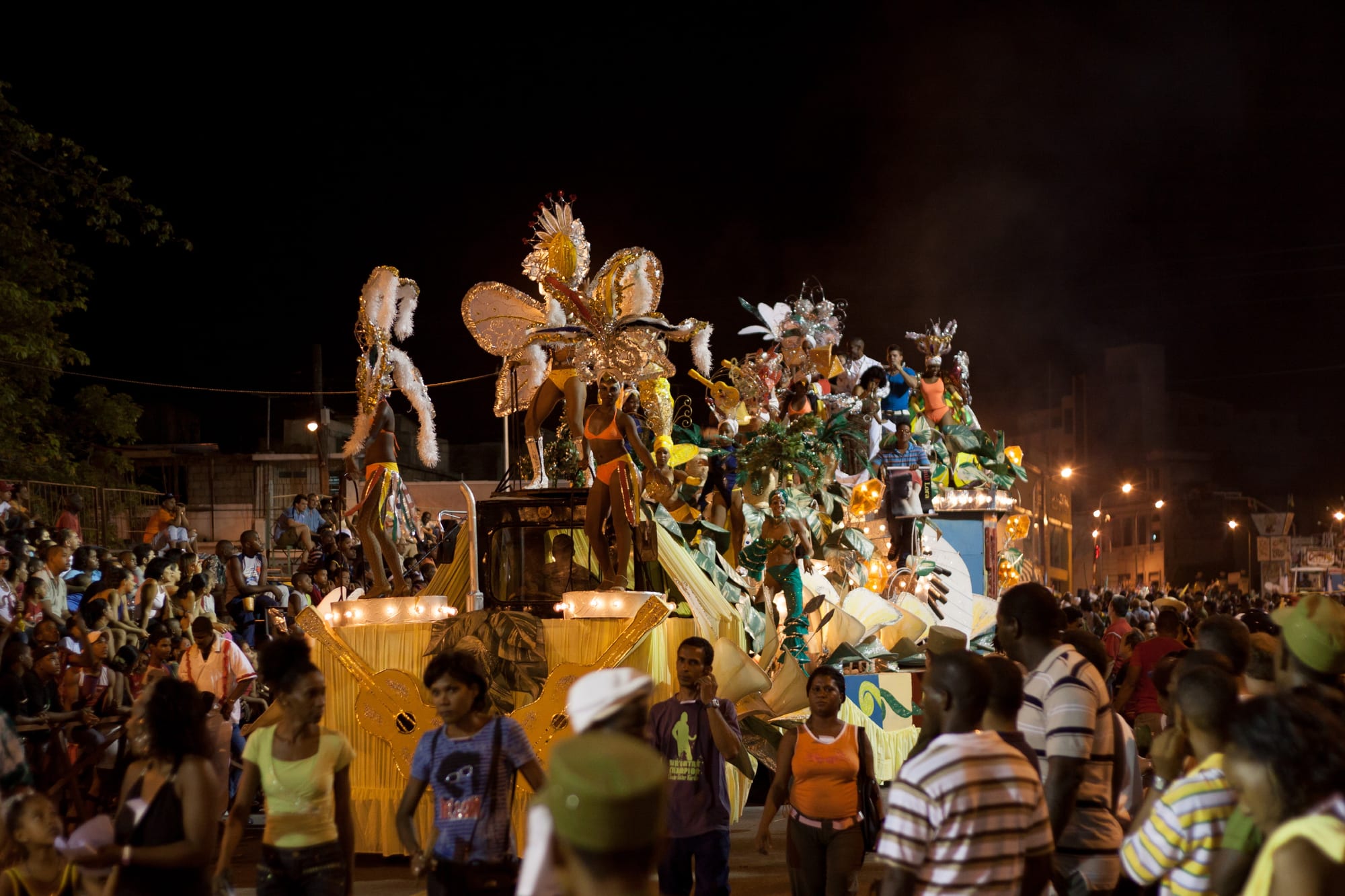 A large illuminated carnival float at night carrying dancers in feathered golden costumes and giant headdresses, moving past a dense crowd of spectators