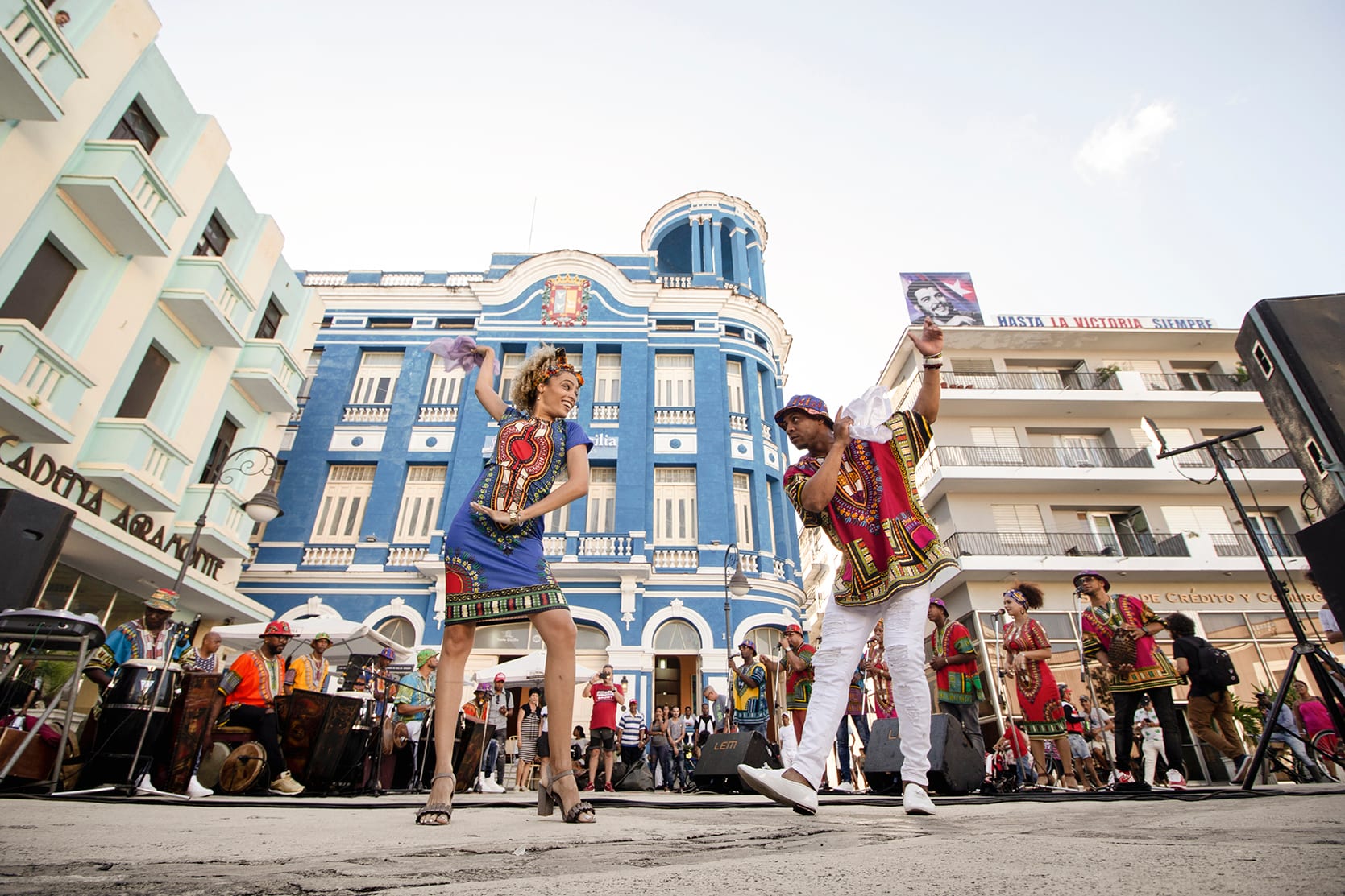 A woman in a blue patterned dress and a man in white trousers and a patterned tunic dance face to face, each holding a white handkerchief aloft, with drummers and a crowd behind them in a square of pastel buildings