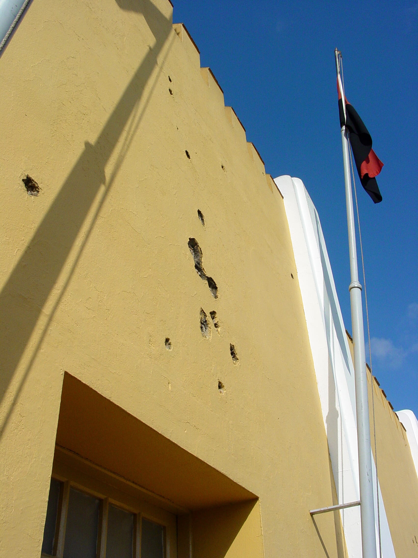 A yellow military barracks building, its walls pocked with bullet-hole markings, now a school