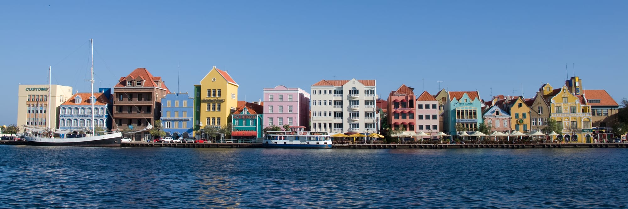 A row of tall, brightly coloured Dutch colonial townhouses along a harbour channel, with a small ferry and sailboat moored in front