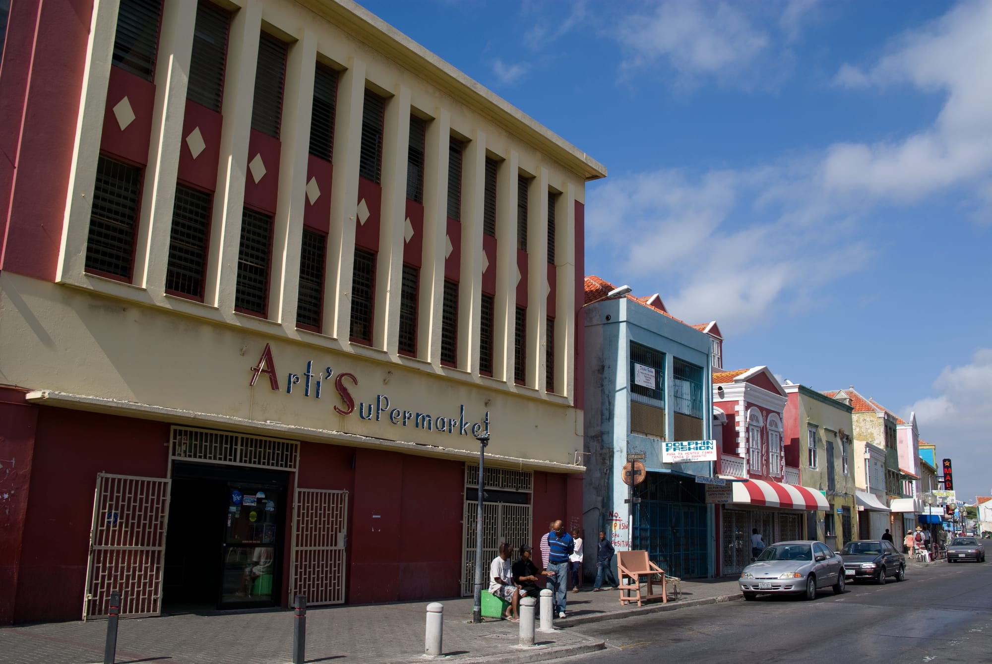 A street of colourfully painted shopfronts including a supermarket, with parked cars and people chatting on the pavement