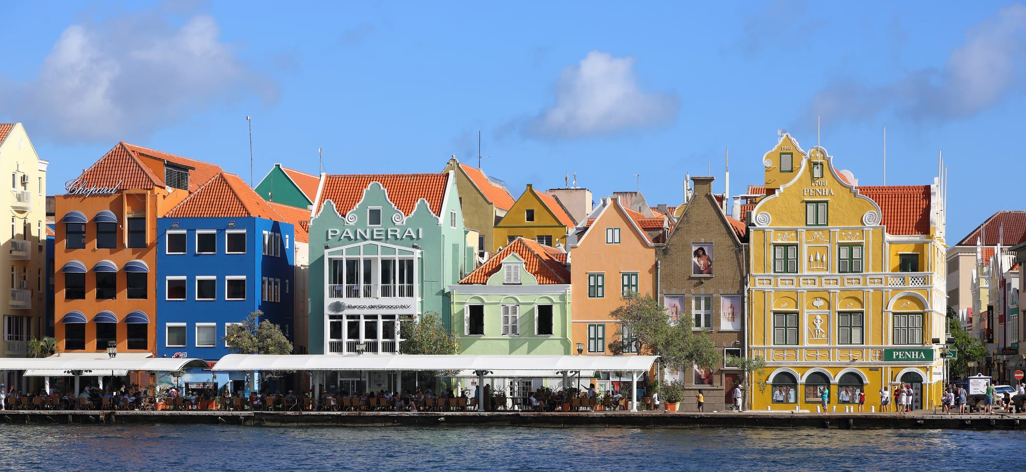 A row of tall Dutch colonial townhouses painted in bright yellows, pinks and blues along a harbour channel
