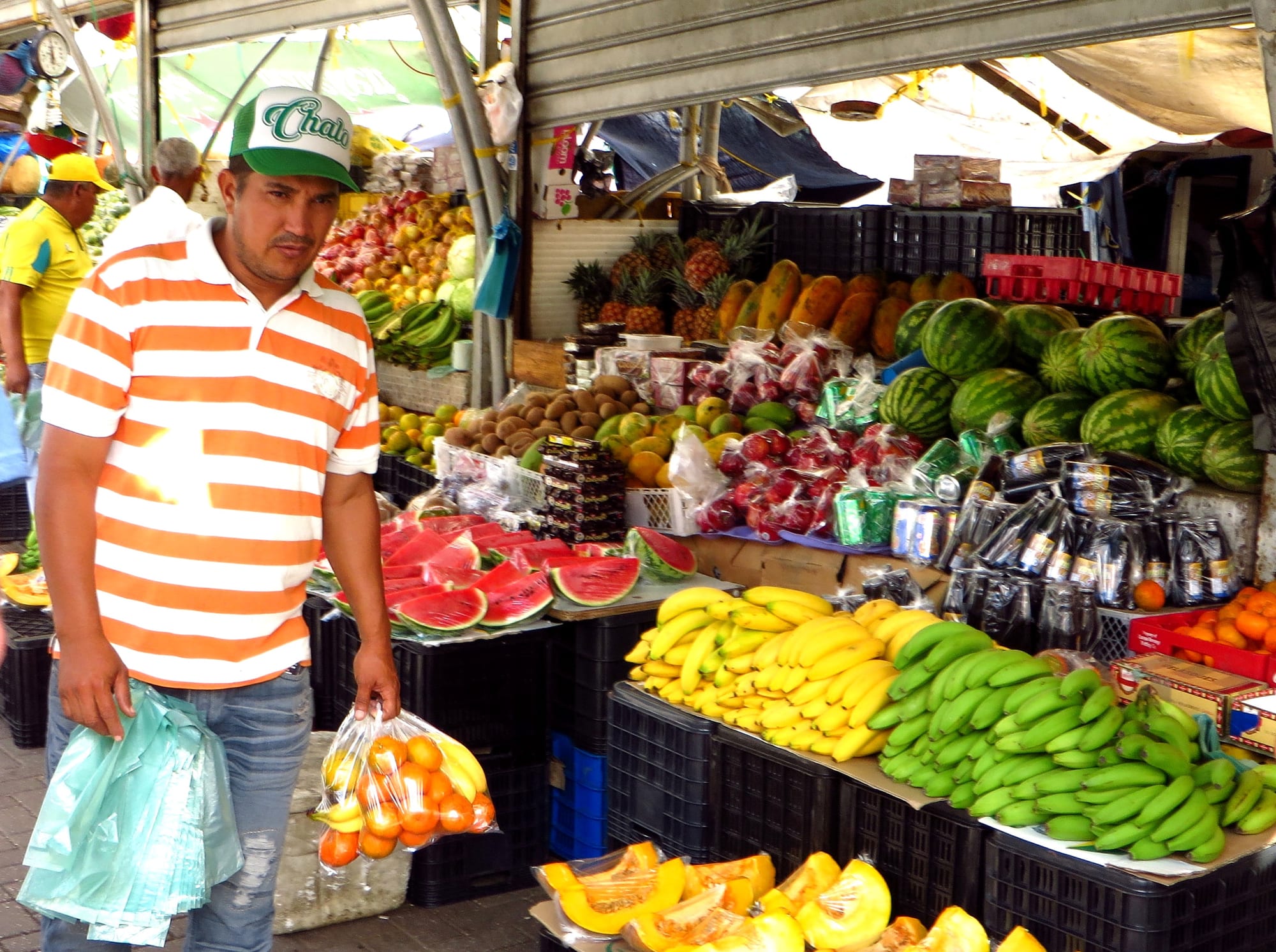 A man carrying bags of fruit past a market stall piled with watermelons, bananas, pineapples and papayas