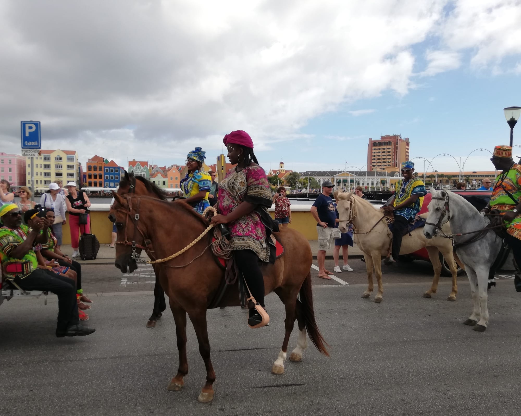 Riders in brightly patterned African-print clothes and head wraps on brown, cream and grey horses in a street, with onlookers and a row of colourful waterfront buildings across the harbour behind