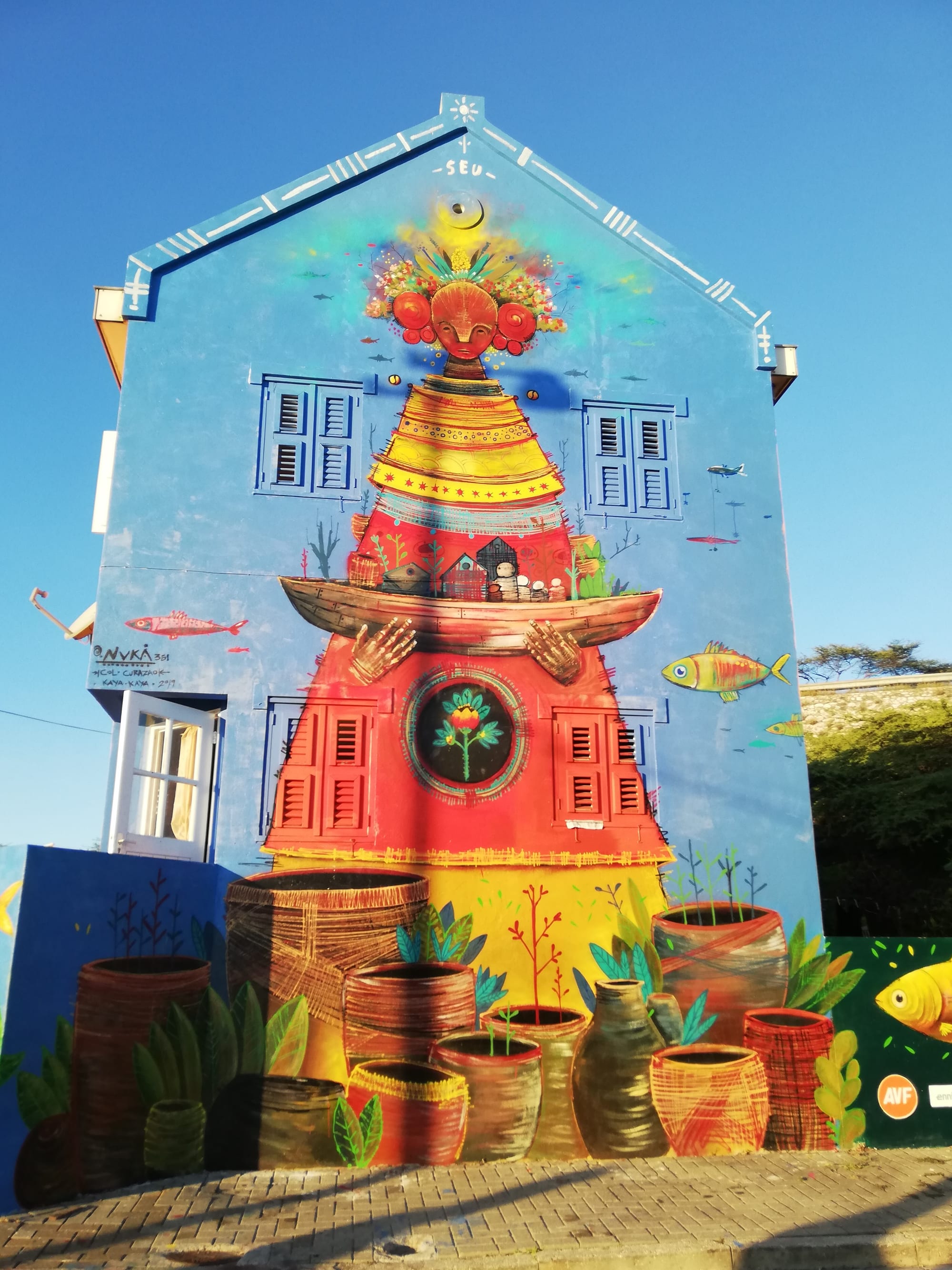 A mural covering the gable end of a blue house, showing a tall figure in a red and yellow dress with a flowered headdress, holding a boat full of houses, with painted woven baskets and pots across the bottom