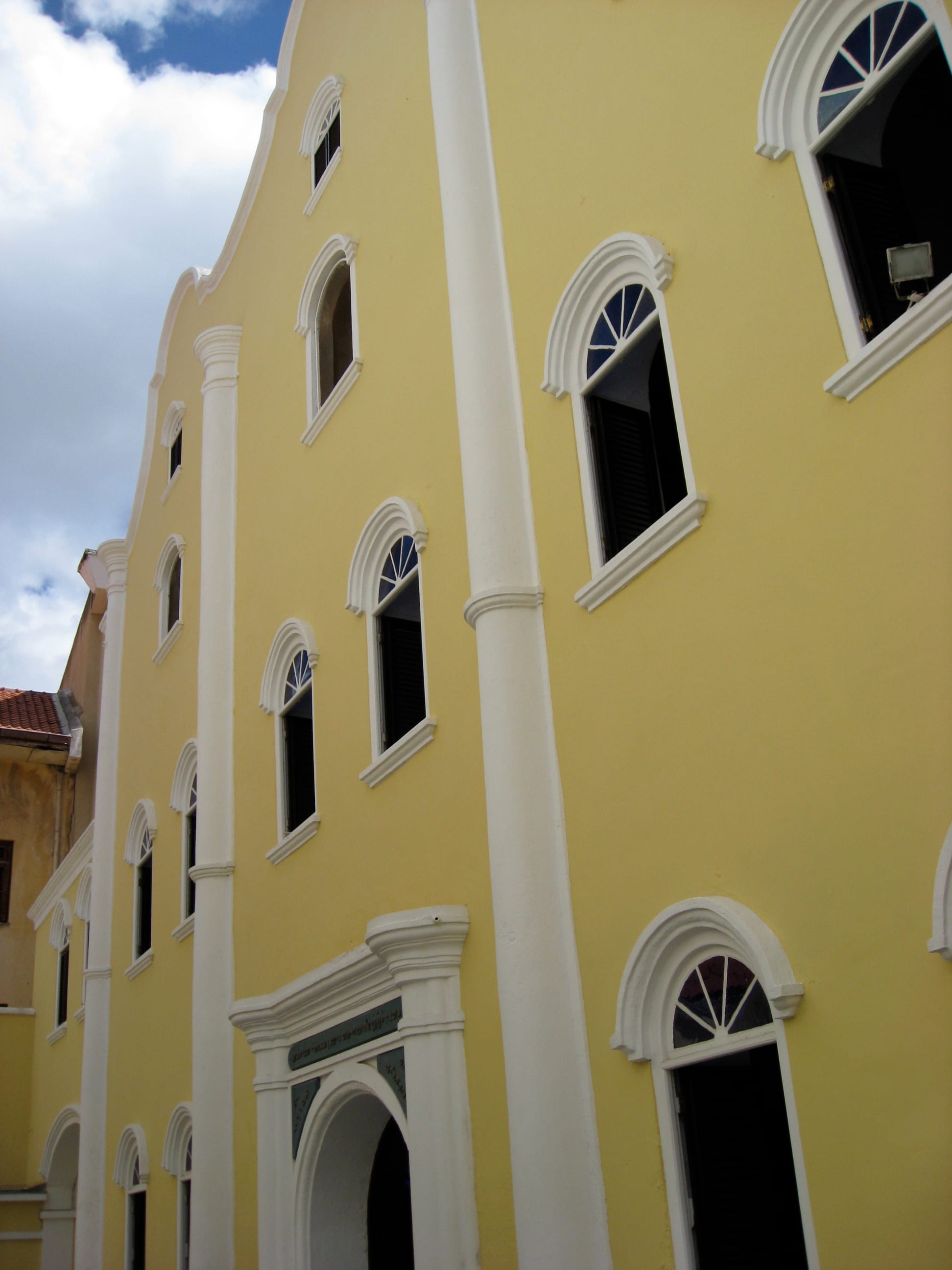 The tall yellow facade of an eighteenth-century synagogue with white pilasters, a curved gable and rows of arched windows with dark shutters