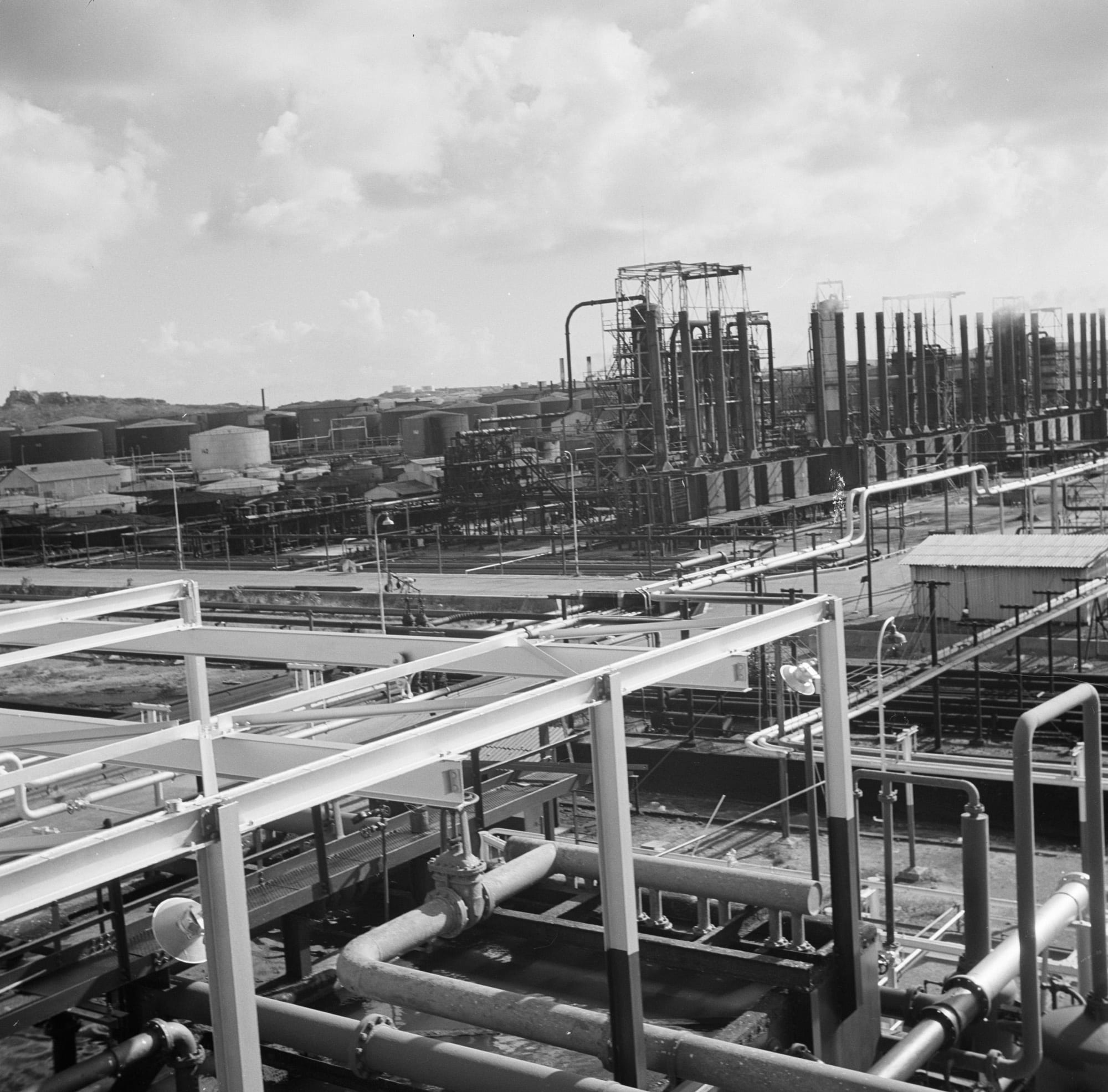 A black-and-white photograph of an oil refinery: pipelines and steel walkways in the foreground, rows of tall stacks and storage tanks behind